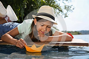 Cute little girl playing with paper boat on wooden pier near river