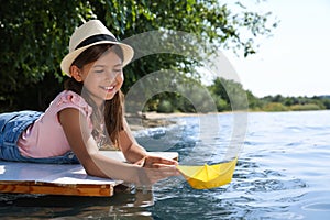 Cute little girl playing with paper boat on wooden pier near river