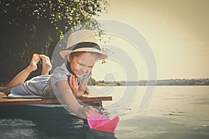 Cute little girl playing with paper boat on wooden pier near river. Retro photo effect