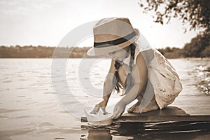 Cute little girl playing with paper boat on wooden pier near river. Retro photo effect