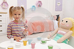 Cute little girl playing tea party with doll at table in room, space for text