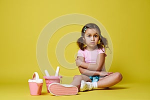 A cute little girl with ponytails sitting in summer clothes with beach toys: a bucket, a watering can and a rake with a shovel