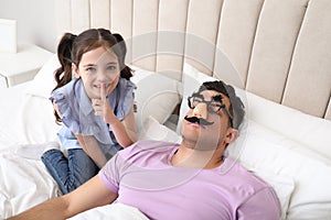 Cute little girl putting funny glasses on father while he sleeping in bed at home