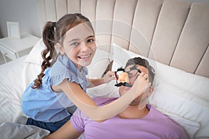 Cute little girl putting funny glasses on father while he sleeping in bed at home