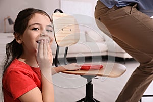 Cute little girl putting whoopee cushion on father`s chair while he sitting down at home, closeup