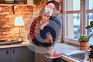 Cute little girl in rabbit makeup and hat sitting on hands of his father in loft style kitchen at morning.