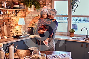 Cute little girl in rabbit makeup and hat sitting on hands of his father in loft style kitchen at morning.