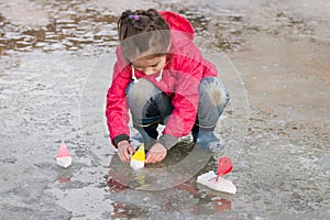 Cute little girl in rain boots playing with ships in the spring water puddle