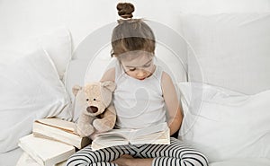 Cute little girl reading a book on the bed in the bedroom