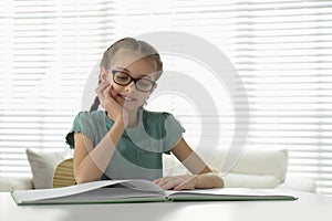 Cute little girl reading book at desk in room. Space for text