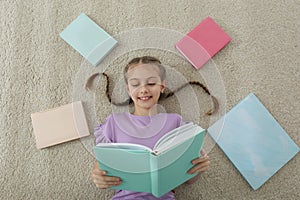 Cute little girl reading book on floor at home, top view