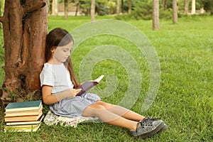 Cute little girl reading book on grass near tree in park