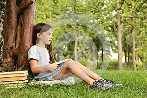 Cute little girl reading book on green grass near tree in park