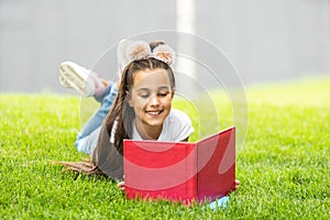 Cute little girl reading book on green grass near tree in park