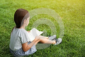 Cute little girl reading book on green grass in park