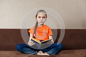 Cute Little Girl Reading a Book  at Home on Sofa Cross Legged. Distance Learning Education. Digital detox