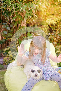 cute little girl reading a book sitting on a pouf with her dog in the garden