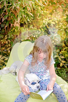 cute little girl reading a book sitting on a pouf with her dog in the garden