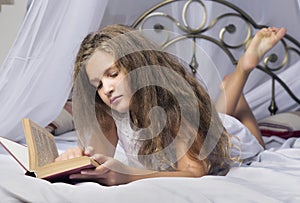 Cute little girl reading a book and smiling while lying on a bed in the room