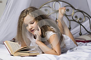 Cute little girl reading a book and smiling while lying on a bed in the room