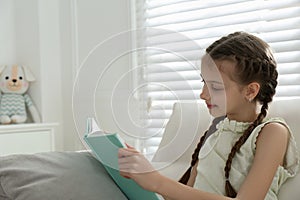 Cute little girl reading book on sofa at home