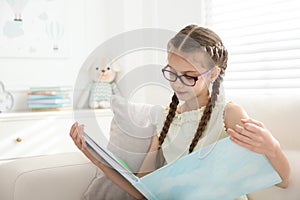 Cute little girl reading book on sofa at home