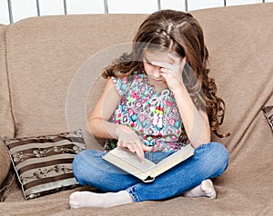 Cute little girl is reading the book on the sofa