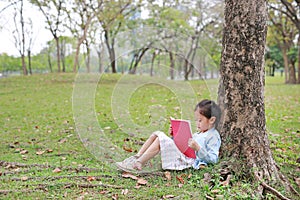 Cute little girl reading book in summer park outdoor lean against tree trunk in the summer garden