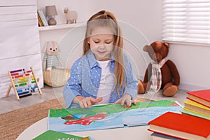 Cute little girl reading book at table in room