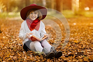 Cute little girl in red hat sitting on fall leaves and reading interesting book