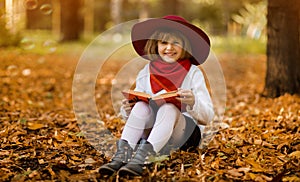 Cute little girl in red hat sitting on fall leaves and reading interesting book