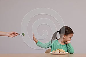 Cute little girl refusing to eat vegetables at table on grey background