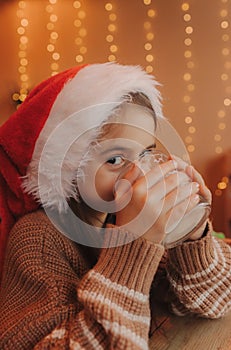 Cute little girl in Santa hat drinks milk from a glass at home. Xmas home holiday celebration