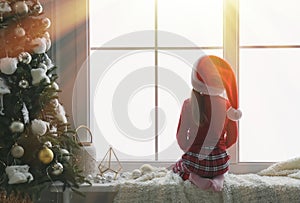 Cute little girl in Santa hat on window sill near Christmas tree at home, back view