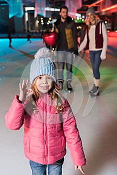 cute little girl showing ok sign and smiling at camera while ice skating with parents