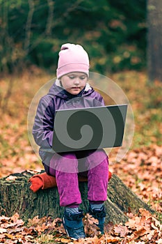 A cute little girl sits on an old stump in the middle of the forest and uses a laptop for online learning or playing games