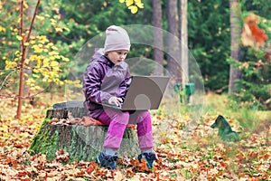 A cute little girl sits on an old stump in the middle of the forest and uses a laptop for online learning or playing games