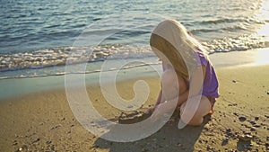 a cute little girl sits on the seashore, playing in the sand and with pebbles.