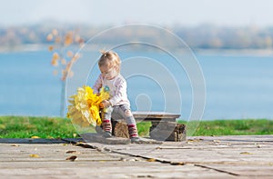 Cute little girl sits on a wood bench with bouquet of maple leaves against the background of an autumn river in park