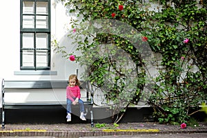 Cute little girl sitting on a bench by giant blossoming rose bush by a house in a typical Amsterdam street, Netherlands