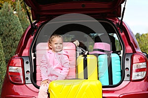 Cute little girl sitting in car trunk loaded with suitcases