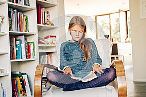 Cute little girl sitting in a chair at home and reading a book