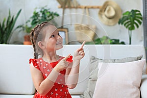 Cute little girl sitting on cozy sofa and pointing up on something intresting by fingers photo
