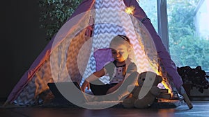 A cute little girl sitting cross-legged on the floor in a pink wigwam watching cartoons on a tablet thoughtfully lifting