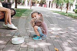 Cute little girl sitting and drawing with chalk on asphalt in park.