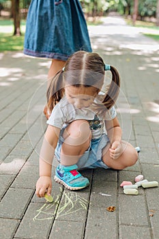 Cute little girl sitting and drawing with chalk on asphalt in park.