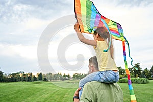 Cute little girl sitting on father`s shoulders and holding colorful kite in the beautiful green park on a warm summer