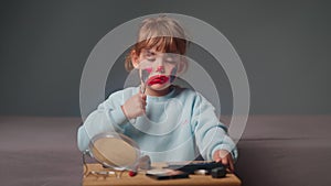 A cute little girl sitting in front of a mirror tries to do her makeup by dipping a brush into cosmetics