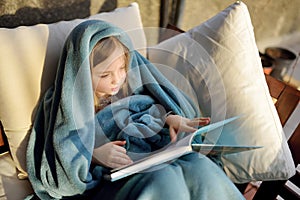 Cute little girl sitting in a garden under a blanket and reading a book on sunny summer evening