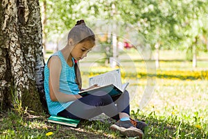 Cute little girl sitting on a green grass in summer park and reading a book